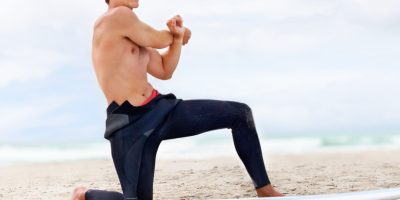 Surfer, man is stretching and beach with surfboard for ocean waves and start fitness, adventure and travel. Nature, vacation and surfing in Hawaii, summer holiday on an island and extreme sports.
